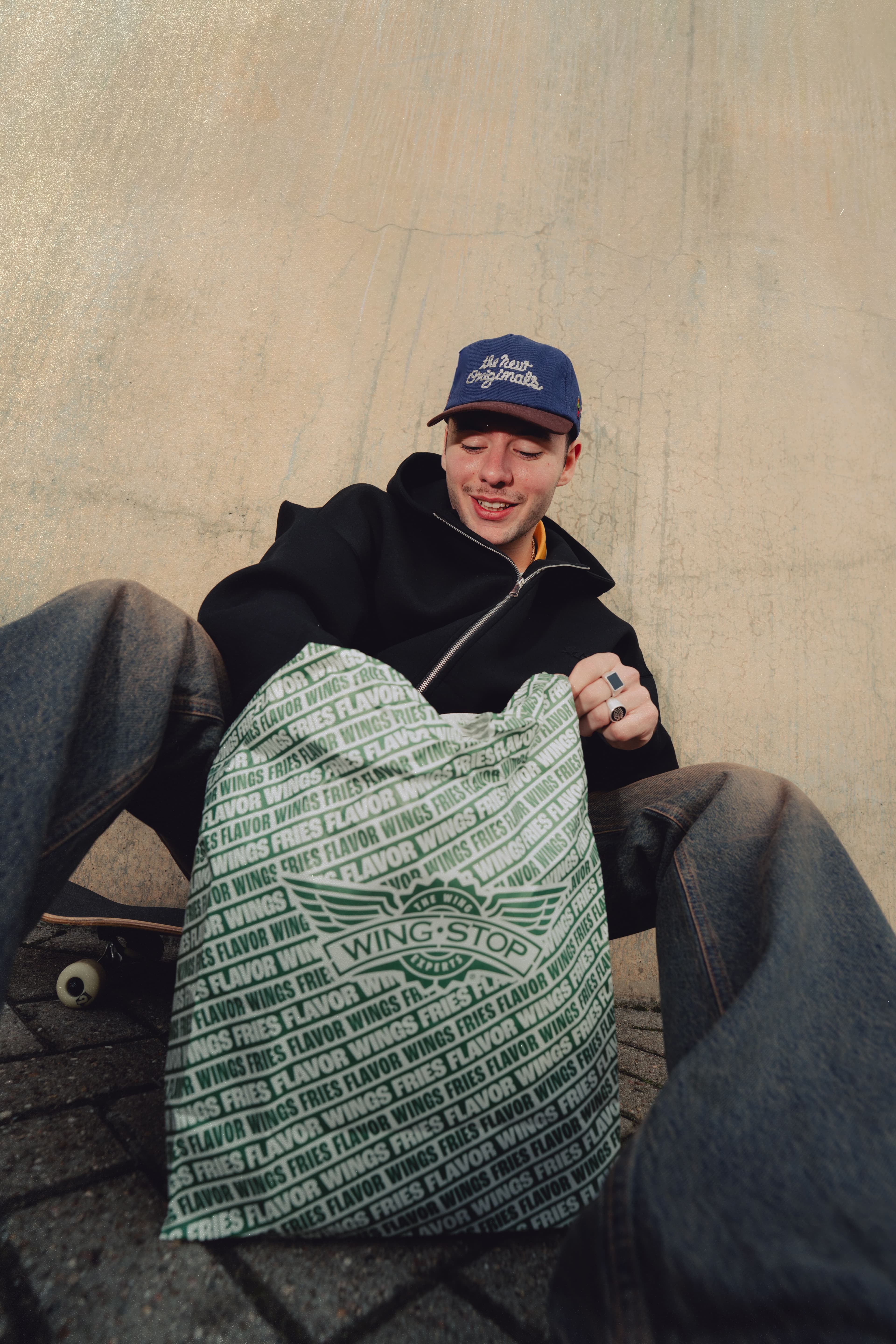 A man sitting next to a brick wall with a hat and bag with Wingstop written on it